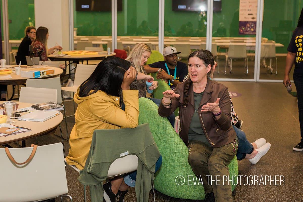 Becky McCray is talking with a woman with dark skin and hair who cradles her head on one hand while thinking.