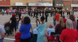 A diverse crowd watches a marching band in a small town parade