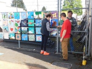 Students hanging art squares from a chain link fence. 