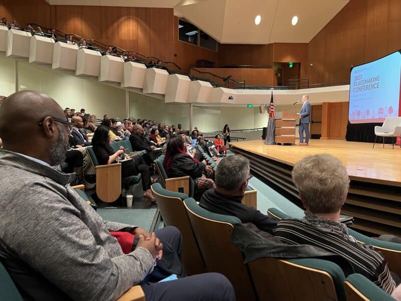 A diverse crowd of people seated in auditorium, listening as a speaker on stage opens the conference. 