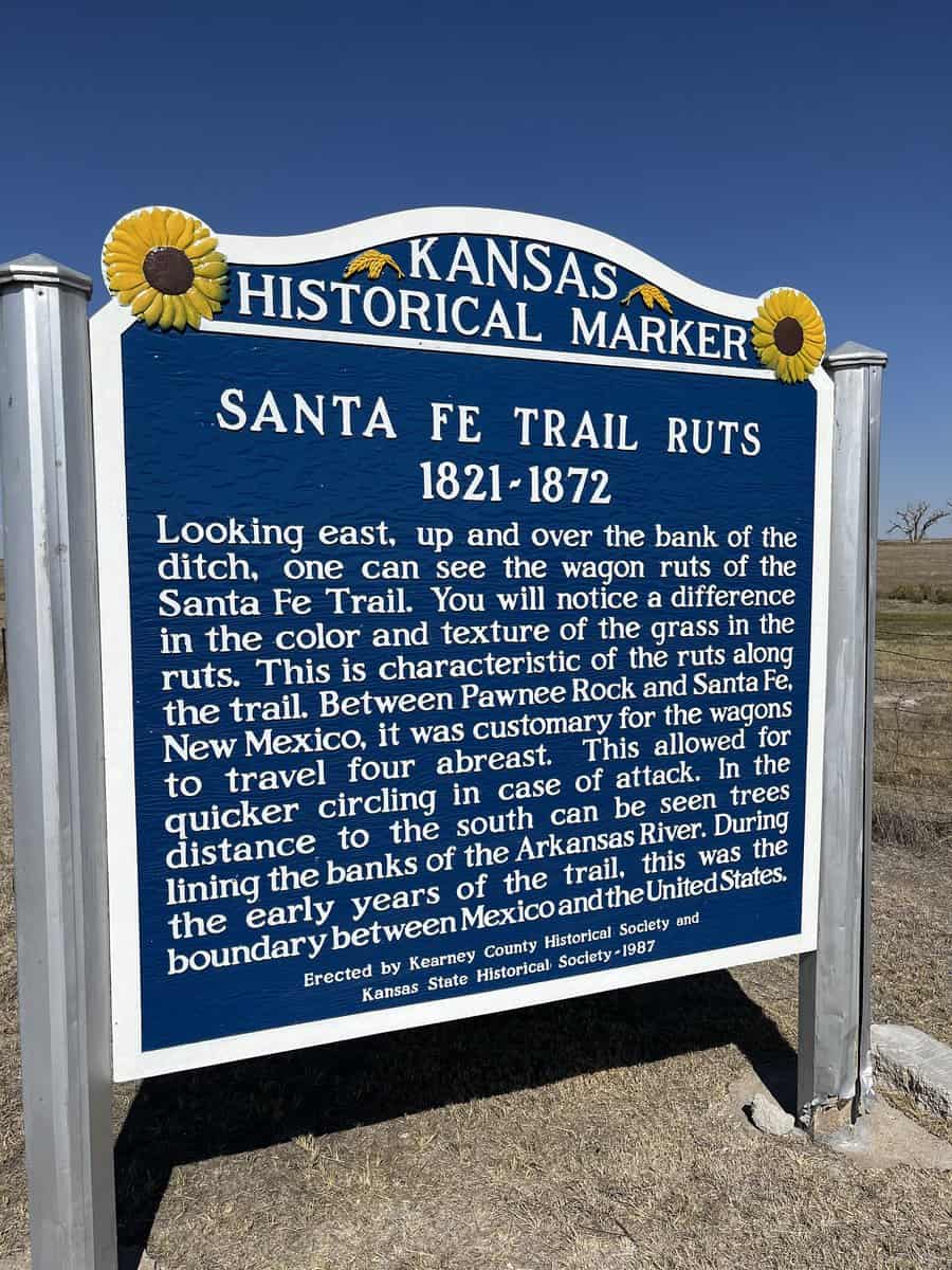Sign text: KANSAS HISTORICAL MARKER SANTA FE TRAIL RUTS 1821-1872 Looking east, up and over the bank of the ditch, one can Santa Fe Trail. see the wagon ruts of the You will notice a difference in the color and texture of the grass in the ruts. This is characteristic of the ruts along the trail. Between Pawnee Rock and Santa Fe, New Mexico, it was customary for the wagons to travel four abreast. This allowed for quicker circling in case of attack. In the distance to the south can be seen trees lining the banks of the Arkansas River. During the early years of the trail, this was the boundary between Mexico and the United States. Erected by Kearney County Historical Society and Kansas State Historical Society-1987