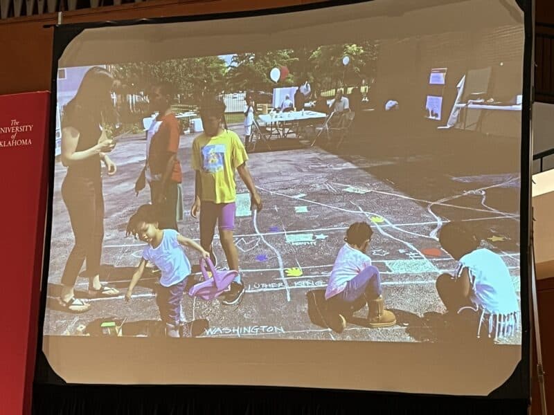 Photo of a slide projected on screen. Children are playing and drawing on a chalked map of their neighborhood. 