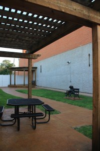 A building with no roof has been converted to a pocket park with a small stage, a sculpture, and a picnic table surrounded by green space.