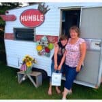 A tiny travel trailer being used as a mobile retail store, two customers are smiling just outside with their purchase.