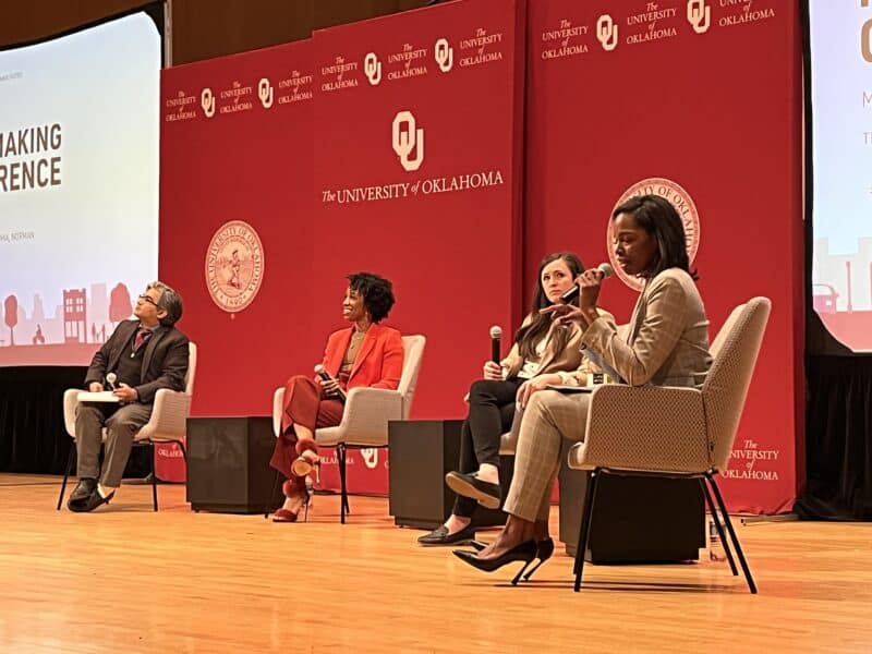 A diverse panel of people seated in chairs on a stage, all holding microphones. One woman is leading the discussion. 